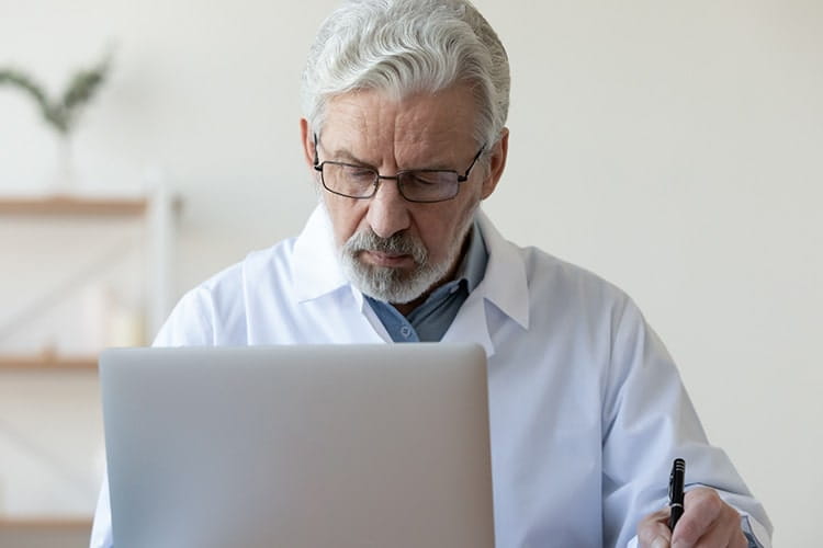 Serious old doctor therapist making notes in medical journal doing online research using laptop in hospital office. Senior adult male physician working on computer writing prescription sits at desk.