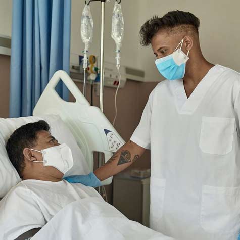 Masked and gloved male nurse reassures a masked male patient in hospital bed. 