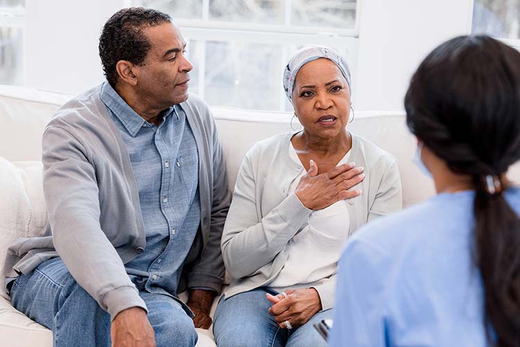 A Black woman wearing a headscarf and touching her heart speaks to a healthcare professional while her husband looks on.