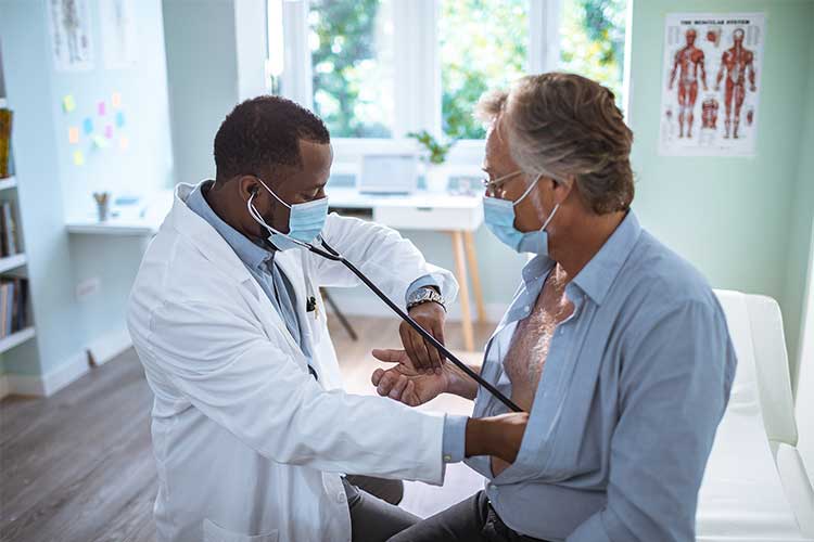 A Black male doctor listens to a white male patient's heart with a stethoscope while simultaneously taking his pulse rate with his other hand. Both people are wearing masks.
