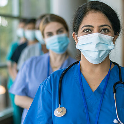 Female healthcare worker wearing a mask, scrubs and a stethoscope smiles into the camera. Behind her are other medical team members wearing masks.