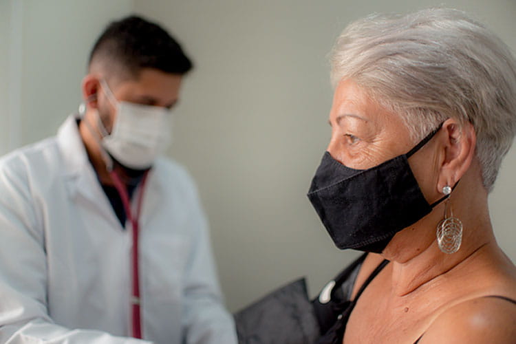 Male healthcare worker wearing a white coat and mask checks the blood pressure of an elderly woman wearing a black mask.