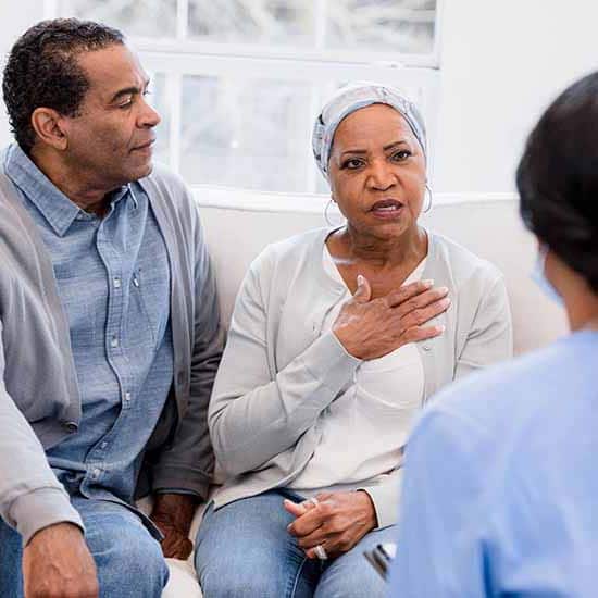 A Black woman wearing a headscarf and touching her heart speaks to a healthcare professional while her husband looks on.