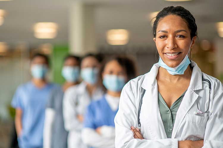 A Black female doctor smiles after pulling her mask down. Behind her are 4 other healthcare workers wearing masks. 