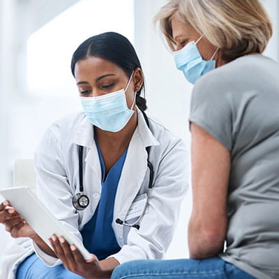 A female healthcare professional of color shares information on a sheet of paper with an older female white patient. Both women are wearing masks. 