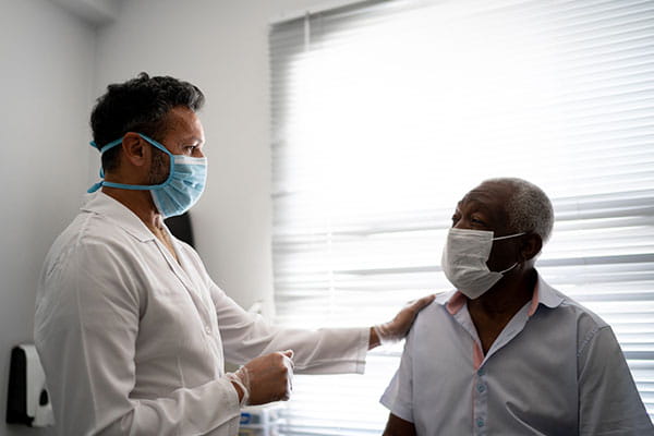 A male healthcare worker reassures his elderly male patient, who is seated in an exam room. Both men are wearing masks.