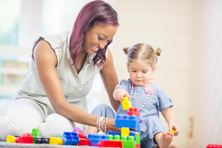 Photo of woman playing with a toddler on the floor. The woman is helping the child build with blocks.