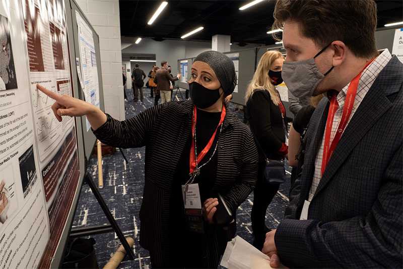 A woman in a mask points at a poster presentation while a man in a mask looks on as she explains. 
