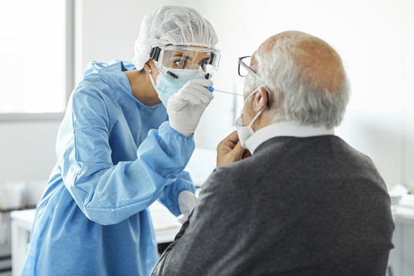 A female healthcare worker in gown, gloves, mask and shield swabs the nose of an elderly man for a COVID-19 test.