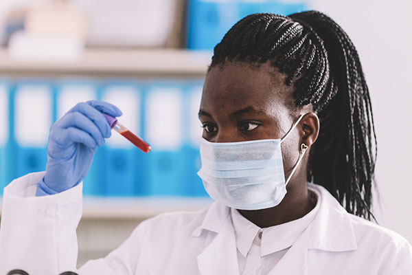 Black female healthcare worker wearing a mask holds up a vial of blood for examination.