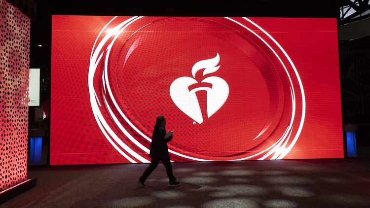 A woman walks in front of a large screen displaying the AHA heart and torch logo inside a circle at Scientific Sessions 2022. 