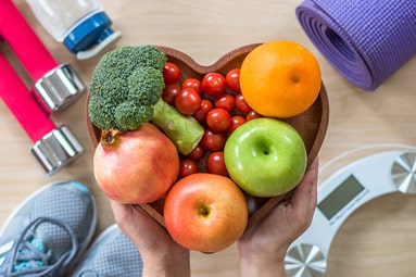 A healthy lifestyle concept showing a small selection of healthy fruits and vegetables in a heart-shaped bowl, a water bottle, sports equipment including barbells, athletic shoes, and a yoga mat, and a scale.scale
