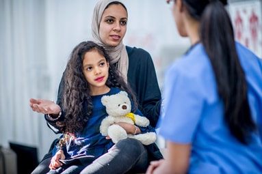 An Ethnic mother and daughter are indoors in a hospital. The girl is holding a teddy bear for comfort. The mother and doctor are talking about the girl's checkup.