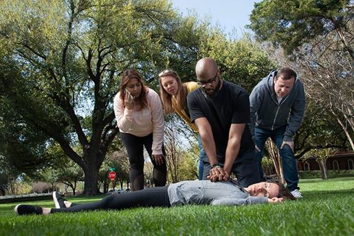 A group of bystanders watch a man perform CPR while another calls 911.