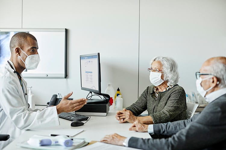 Doctor discussing with senior couple during COVID-19. Male medical professional is explaining elderly man and woman while sitting at desk in clinic. They are wearing protective face masks.