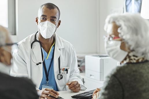 Doctor discussing with elderly couple during COVID-19 outbreak. Male healthcare worker is explaining senior man and woman while sitting at desk in clinic. They are in protective face masks.