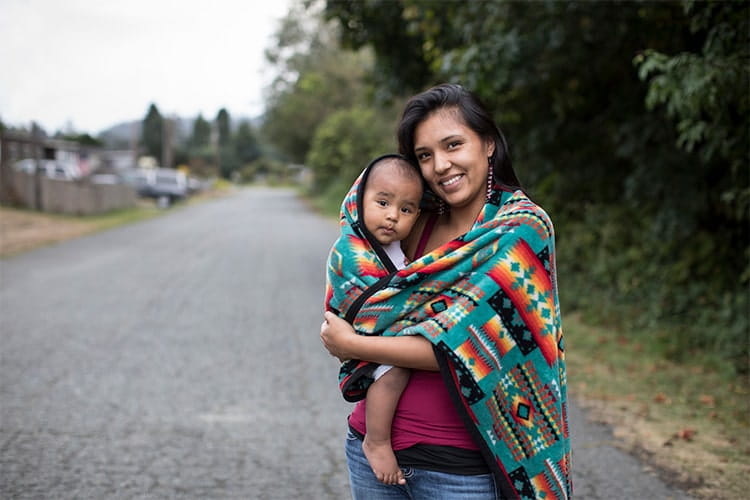 Young Yurok mother walks around her neighborhood, carrying her baby on her hip. They pause to say hello. The baby is barefoot and has an inquisitive expression. The mom wears teeshirt and jeans, and has a blanket with tribal designs wrapped around her baby and herself to stay warm. Houses and trees are soft focus in the background.