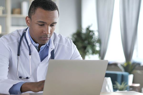 Black male doctor wearing white coat and stethoscope works on laptop computer.