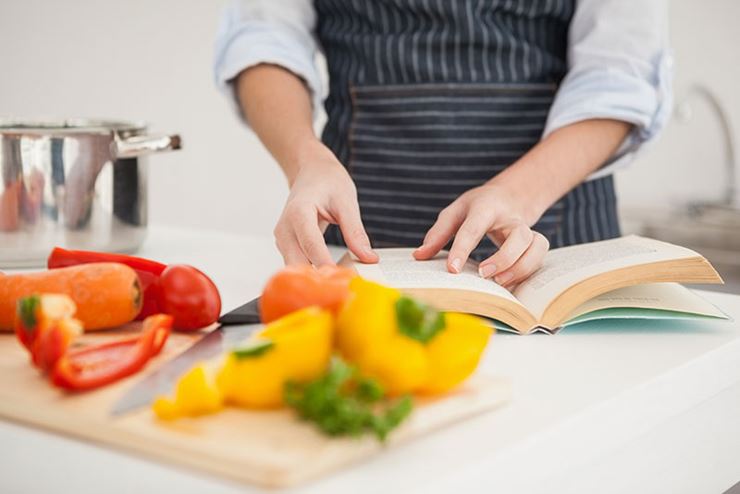 Hands opening cook book with bell peppers, carrots and pot in background