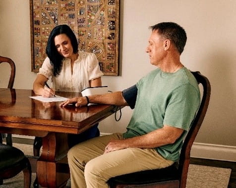 A middle age man sitting at a table having his blood pressure measured. The woman with him is recording his numbers on a form.