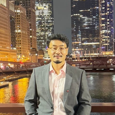 Dr Dai dressed in a shirt and suit jacket, standing in front of a nighttime cityscape
