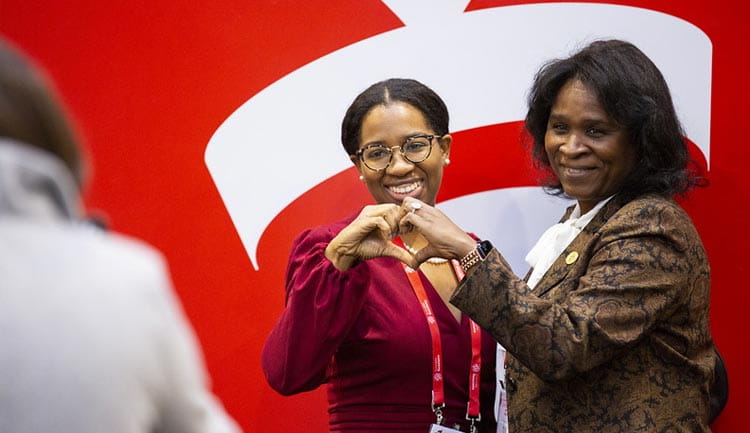 Two attendees at #AHA23 in Philadelphia pose in front of the AHA logo, while creating a heart shape with their hands.