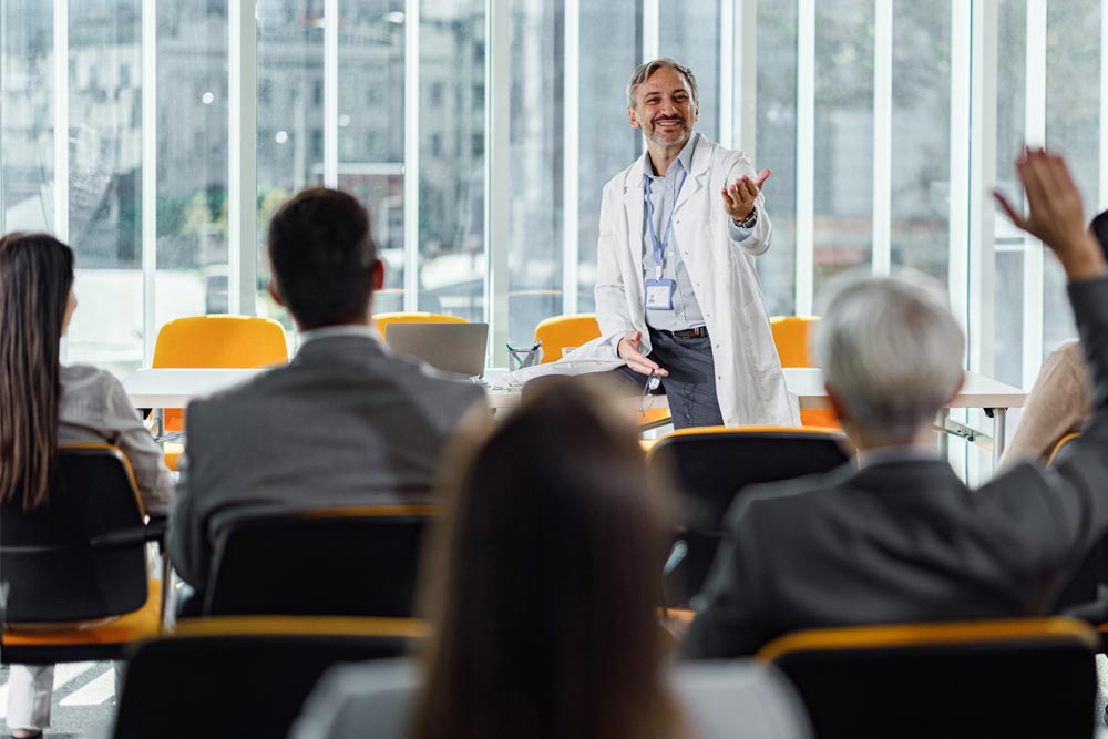 A photo of a man in a white lab coat answering questions from a person in the audience after his presentation.